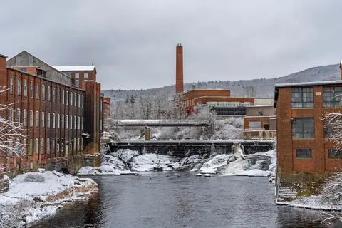 Snow on the brick mill buildings lining both banks of the Millers River, with a falls and a wooded hill behind them.