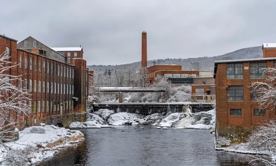 Red brick mill buildings on both banks of a snowy river, with a low dam and a tall smokestack against wooded hills.